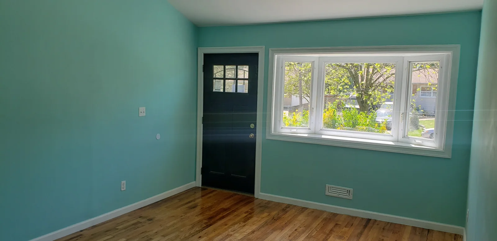 Finished rental property living room with teal walls, white trim, black entry door, large window, and refinished hardwood floor