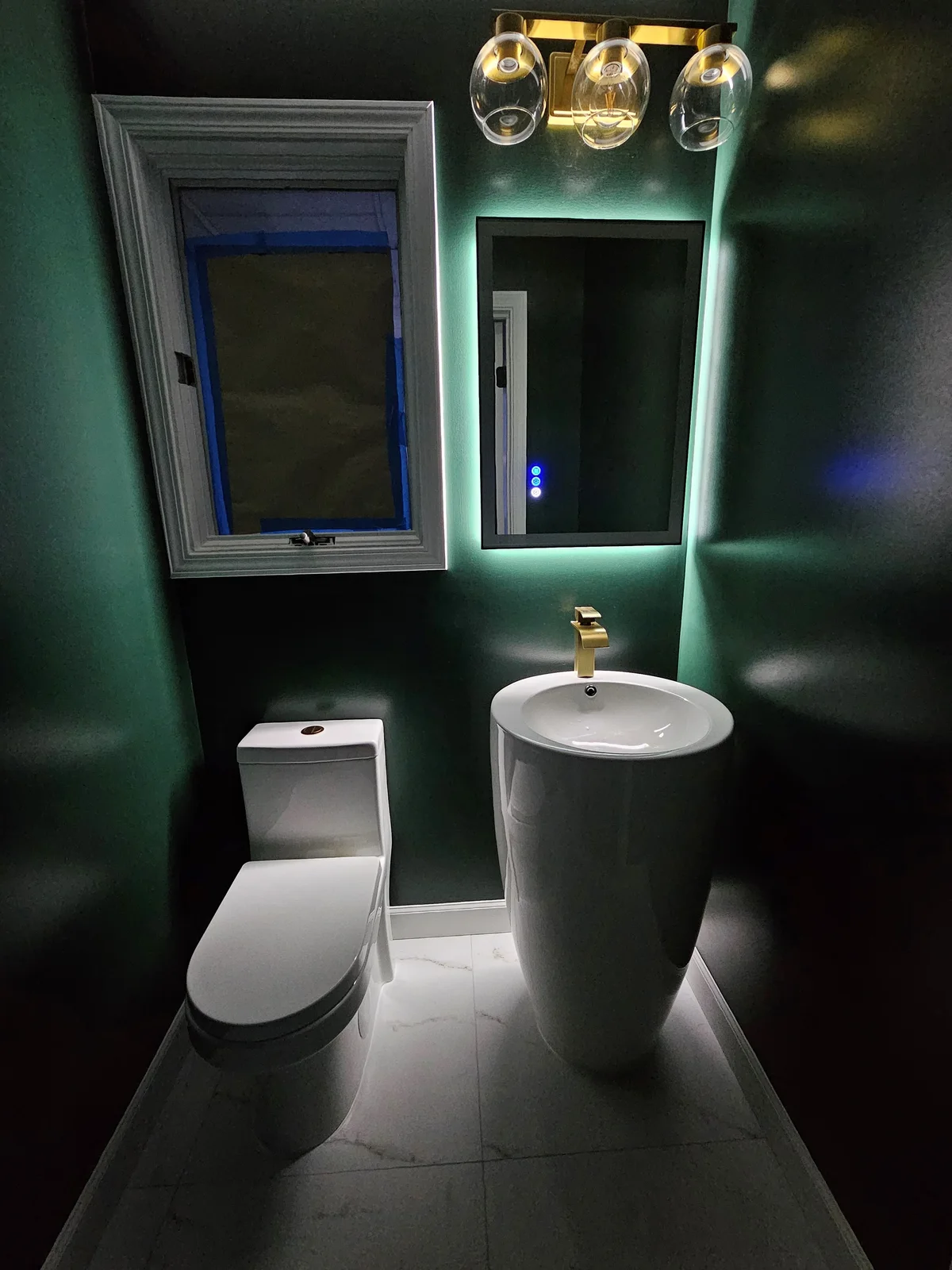 Finished Evansville half-bath remodel with dark green walls, pedestal sink, gold faucet, backlit mirror, and new toilet